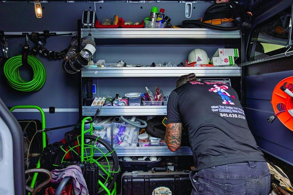 Man in a service van reaches for supplies on shelves. Tools, equipment, and a green hose are visible.