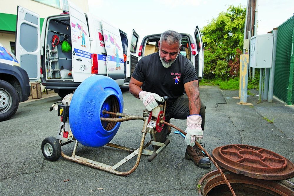 Plumber using drain cleaning machine near open sewer, vans, and metal manhole cover.