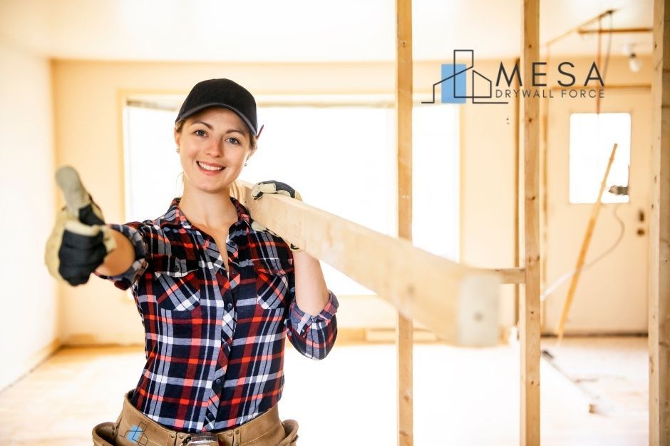 A drywall repair company worker in a plaid shirt and black cap smiles while holding wood in a commercial construction space with wooden framing and bright interior lighting visible near Kloss Ave, and Hill St, Dewey–Humboldt, AZ.