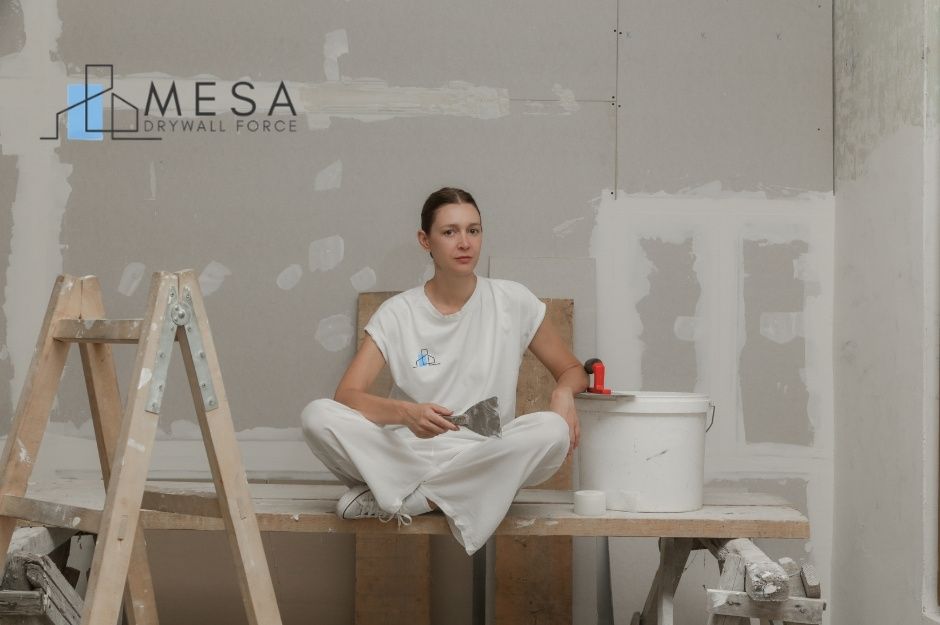 A drywall repair company worker sits cross-legged on wooden sawhorses at a residential construction site, holding a putty knife. She wears white work clothes, surrounded by ladders and joint compound buckets near S 45th Ave, and S Reidar Rd, Laveen, AZ.