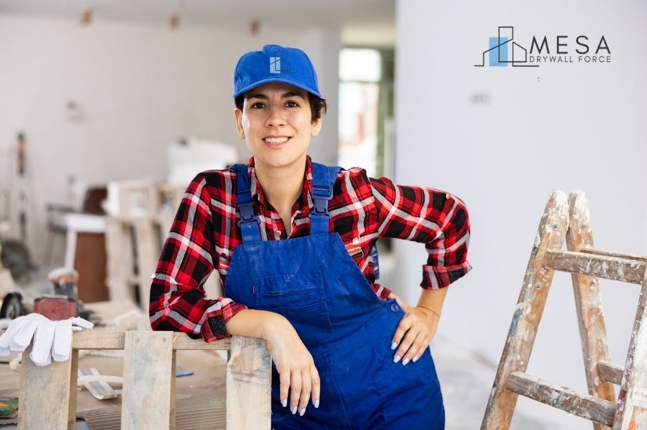 A female drywall repair company worker smiles at a commercial construction site, wearing blue overalls, red plaid shirt, and blue cap. A wooden stepladder is visible nearby, along with work gloves and tools in the interior with unfinished walls near W Aldea Rd, and W Aldea Dr S, Litchfield Park, AZ.