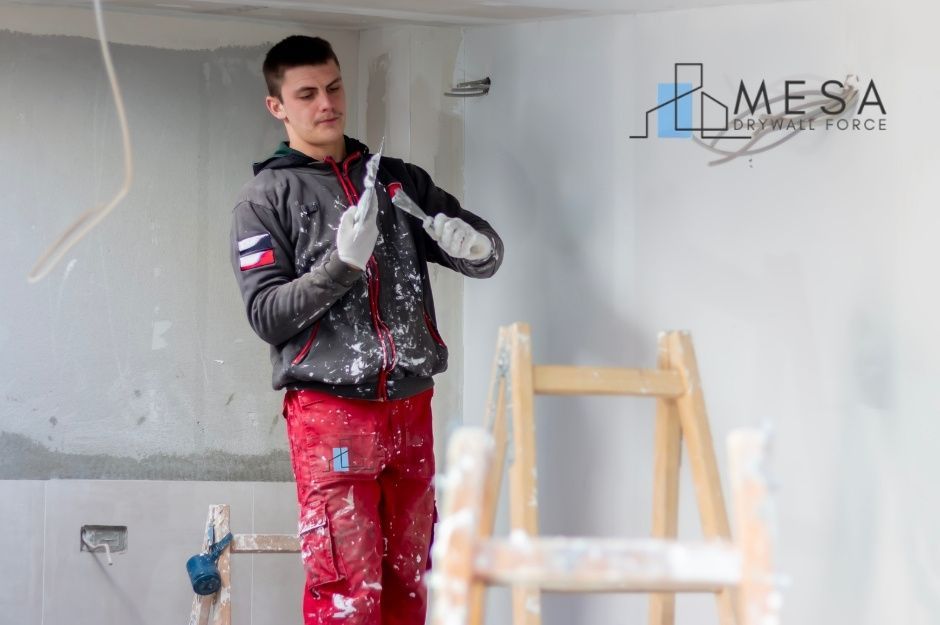 A drywall repair company worker wearing paint-splattered red work pants and a dark gray jacket smooths joint compound using two drywall knives in a commercial room under renovation, with a wooden ladder and unfinished white walls visible near E Lavitt Ln, and N 3rd St in New River, AZ.