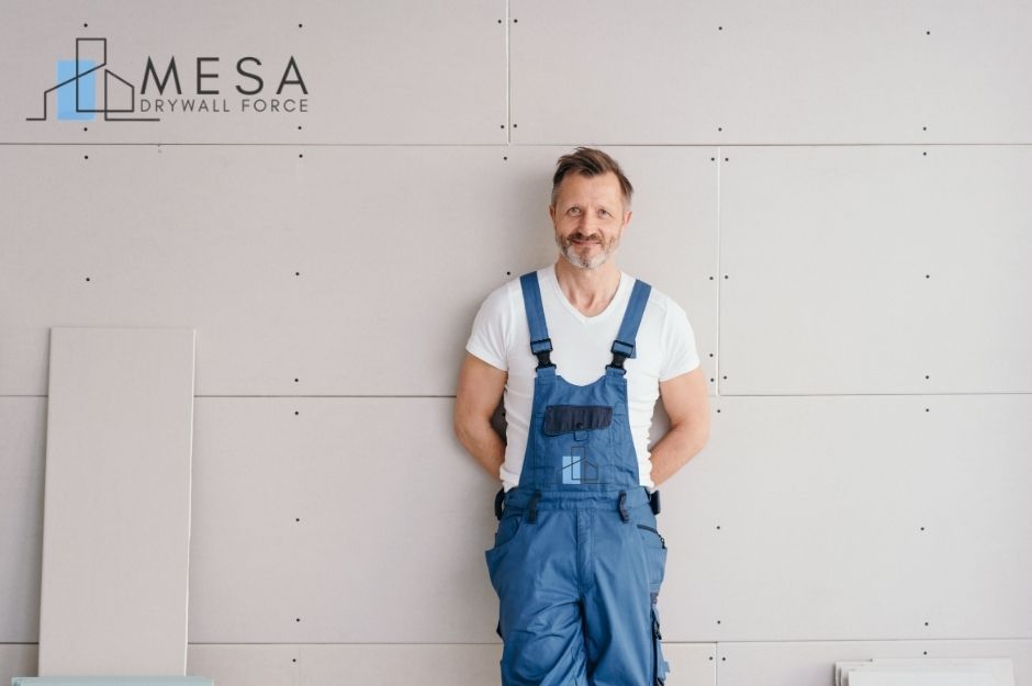 A drywall repair company worker stands in front of a newly installed drywall inside a residential home. He is wearing blue overalls and a white shirt, with his hands behind his back. Several drywall panels are stacked near the wall. Located near E Excelsior Ave, and 209th St, Queen Creek, AZ.