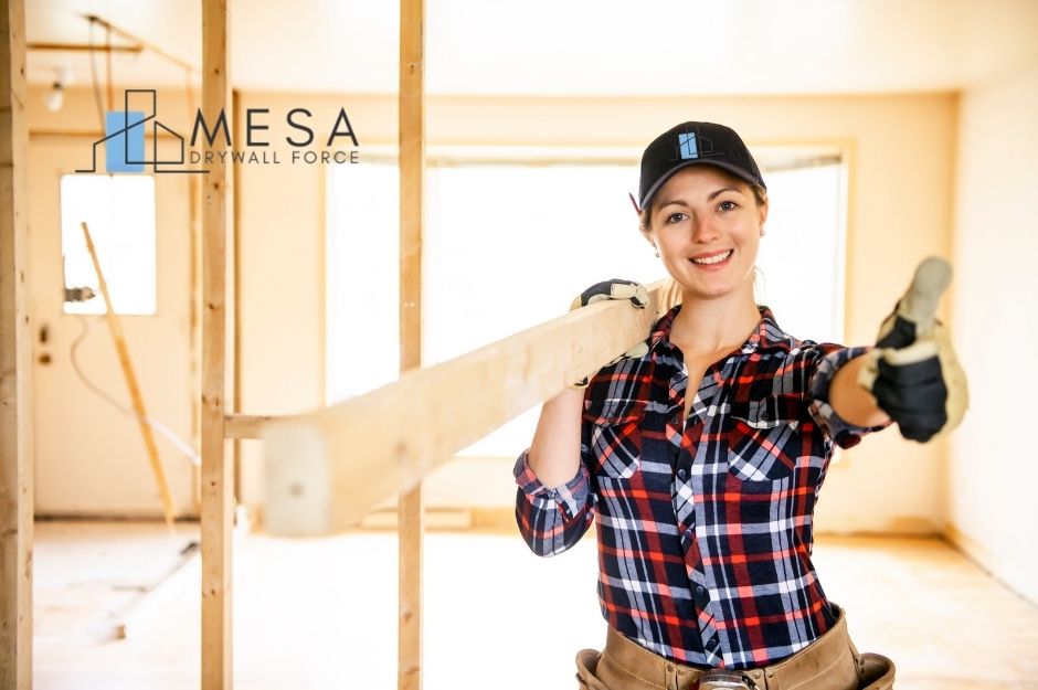 A drywall repair company worker in plaid shirt and black cap carries a wooden board over her shoulder while standing in a commercial construction site with wooden framing and windows visible in the background near N 114th Dr, and W Missouri Ave in Youngtown, AZ.