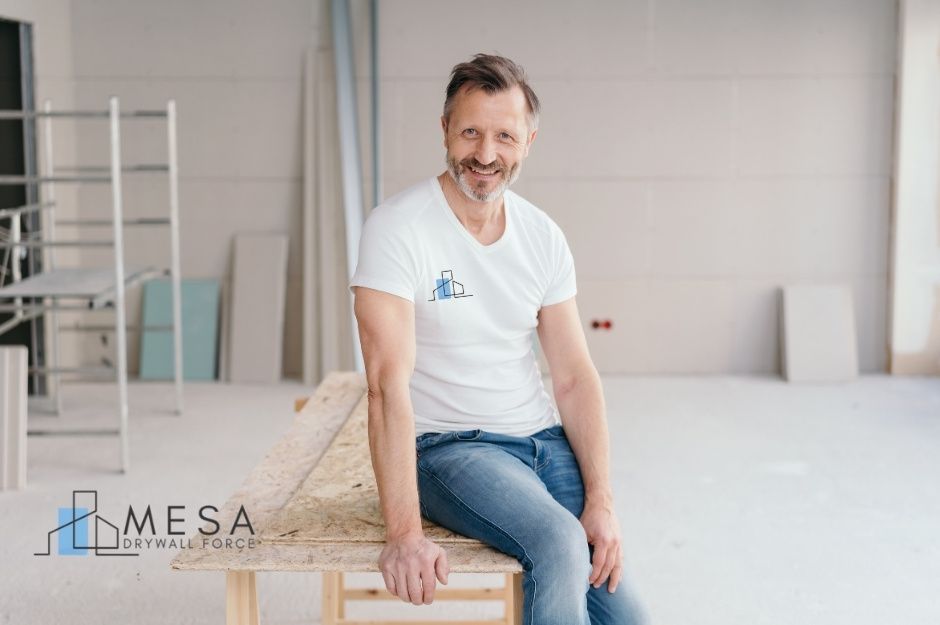 A drywall repair contractor sitting on a wooden workbench inside a partially finished residential room. He is smiling, wearing a white T-shirt and jeans, with construction materials and scaffolding visible behind him. Located near E 39th Ave, and S Descanso Rd, Apache Junction, AZ.
