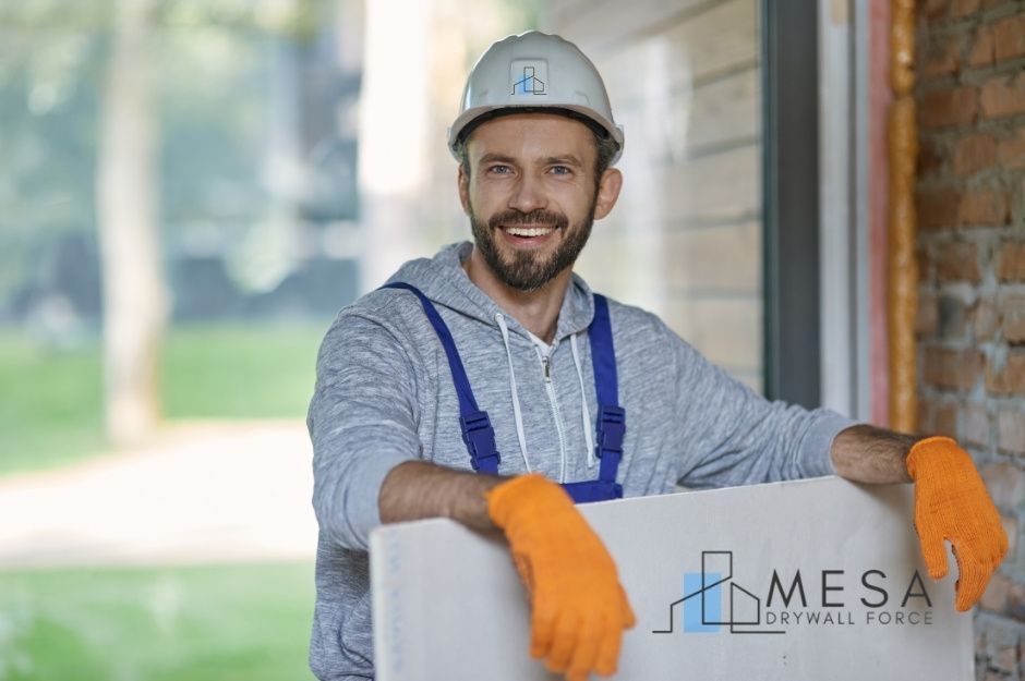 A drywall repair contractor with a beard wearing a white hard hat, gray hoodie, blue overalls, and orange work gloves. He is smiling at the camera and standing next to white drywall sheets in an unfinished residential interior. Located near 9th St, and W Patton Ave, Coolidge, AZ.