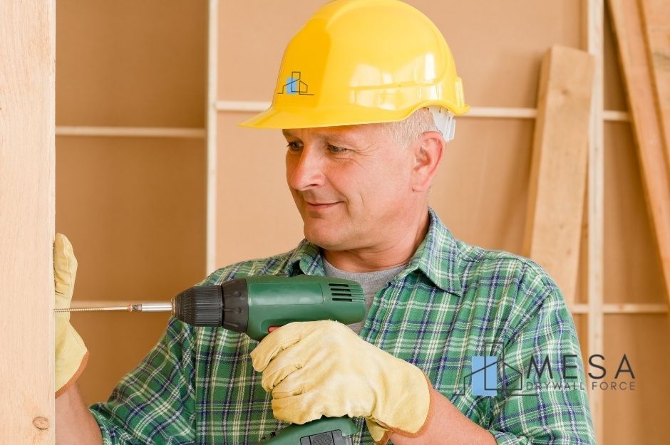 A drywall repair contractor wearing a yellow hard hat and plaid shirt holds a green power drill while working on a residential construction project. He is wearing work gloves and positioned near wooden framing materials. Located near S Cordes Lakes Dr, and E Stagecoach Trail, Cordes Lakes, AZ.