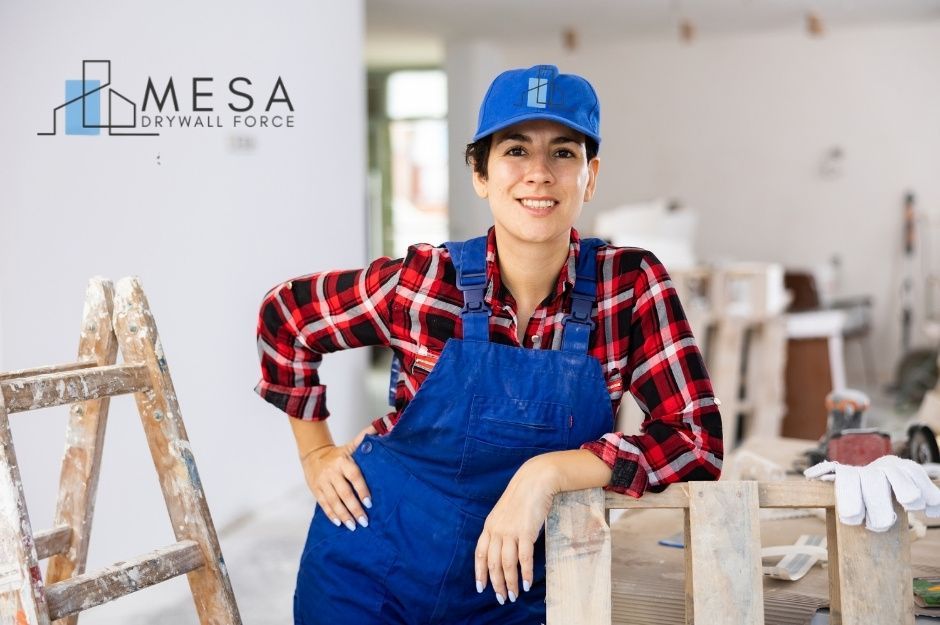 A smiling drywall contractor in blue overalls and a red plaid shirt stands beside wooden construction materials in an unfinished residential room with white walls. A wooden stepladder is visible nearby, along with work gloves and tools. Located near N Turning Leaf Dr, and N Sable Way, Prescott Valley, AZ.