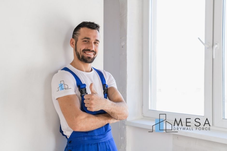 A drywall repair contractor stands indoors wearing blue overalls and a white shirt, smiling confidently while giving a thumbs-up beside a freshly finished white wall and a large window letting in bright light. This is a commercial project located near E Don Carlos Ave, and S McClintock Dr, Tempe, AZ.