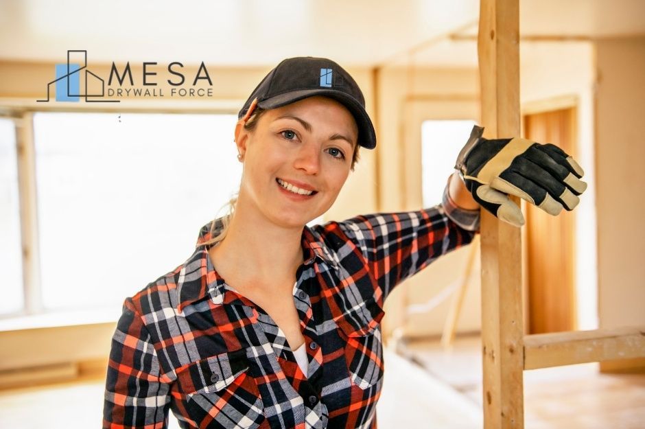 A drywall repair handyman at a residential construction site. A smiling woman in a black cap and orange plaid flannel shirt, wearing work gloves, stands near wooden framing ready for renovation work near W Princeville Ct, and N Crooked Stick Rd, Anthem, AZ.
