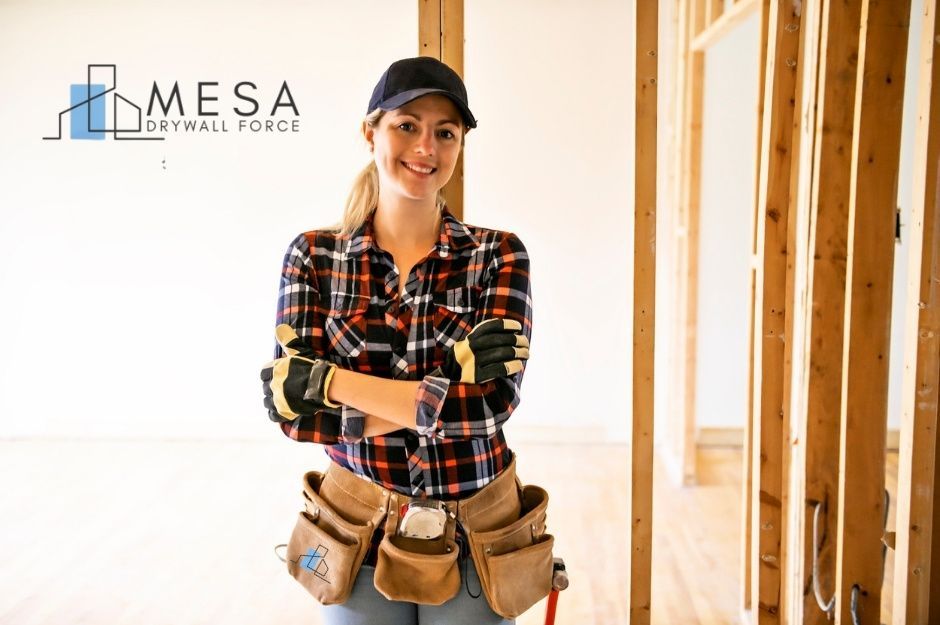 A drywall repair handyman in plaid shirt, black cap, and work gloves stands with arms crossed wearing a tool belt in a residential construction site with wooden framing visible in the background near W Maui Ln, and W Acapulco Ln in El Mirage, AZ.