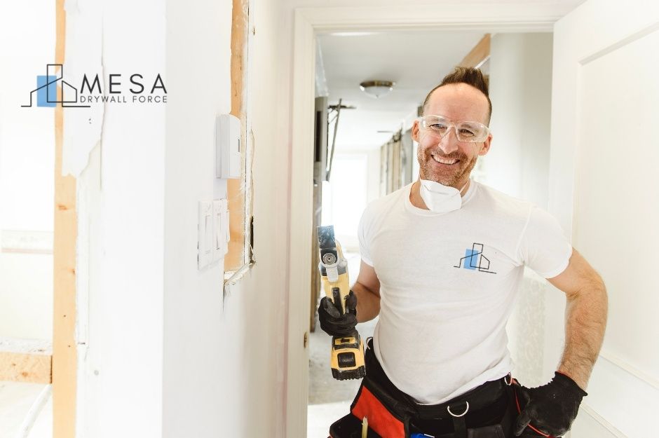 A drywall repair handyman smiling inside a residential hallway, wearing safety glasses, gloves, and a white shirt. He holds a yellow oscillating multi-tool in one hand and has a tool belt around his waist. The wall beside him shows drywall repair in progress near E Aloe Dr, and E Bursage Dr, Gold Canyon, AZ.