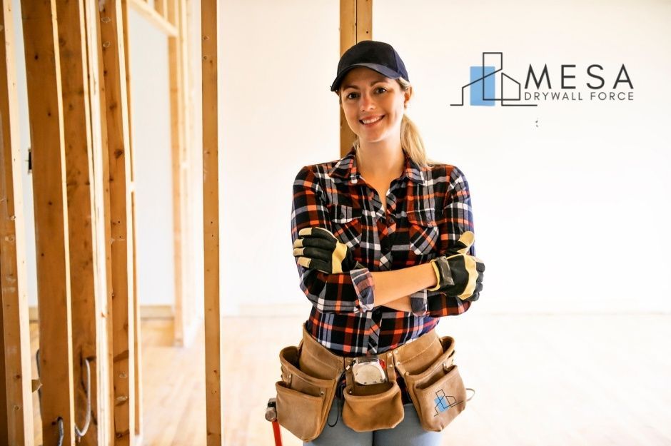 A drywall repair handyman at a commercial construction site, wearing a plaid shirt, black cap, and tool belt, stands with arms crossed in a framed wooden interior near S Lone Spruce Dr, and S Hereford Dr, Mayer, AZ.
