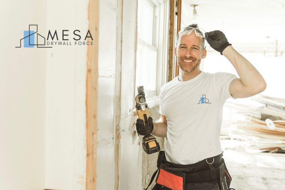A drywall repair specialist smiles while holding a cordless drill, wearing safety glasses, a white t-shirt, and black work pants with a tool belt. He stands in a bright residential construction area with white drywall and wooden framing visible near S 46th St, and E Desert Wind Dr, Ahwatukee Foothills, AZ.