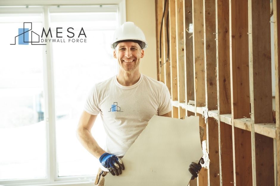A drywall repair specialist smiles while holding a large sheet of drywall in a residential construction site. He wears a white hard hat, a white t-shirt, and blue work gloves. Exposed wooden wall studs are visible behind him with natural light from windows near 20th St, and Maricopa Rd, Florence, AZ.