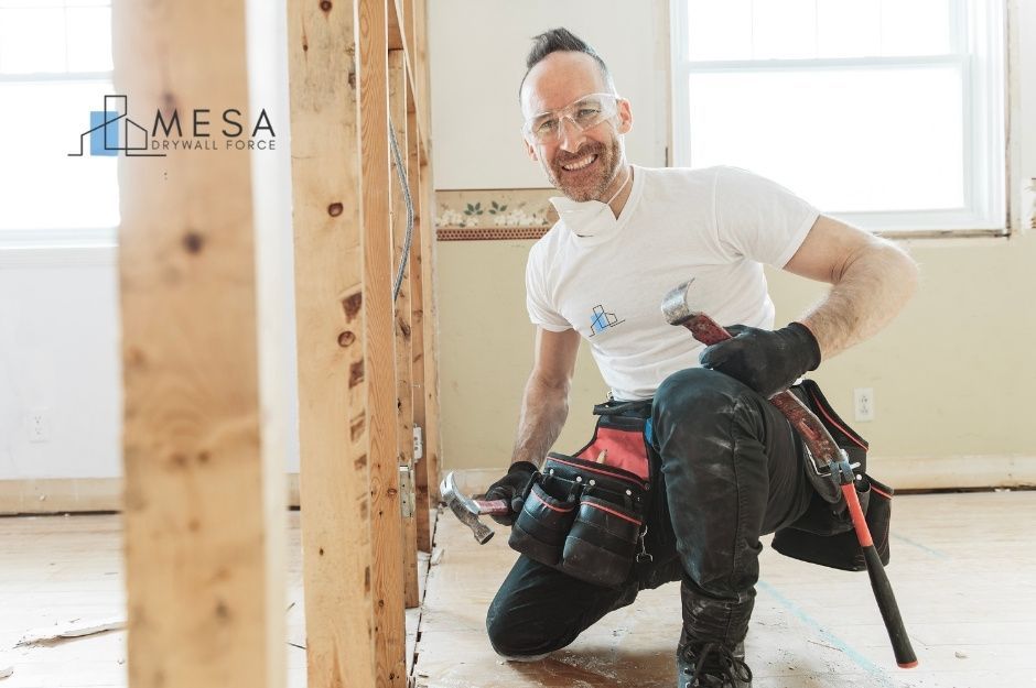 A drywall repair specialist kneeling inside a partially renovated residential room, wearing safety glasses, gloves, and a tool belt. He holds a hammer in one hand and a pry bar in the other, smiling beside exposed wall studs and electrical wiring. Located near E Bellerive Dr, and S Stuart Ave, Gilbert, AZ.