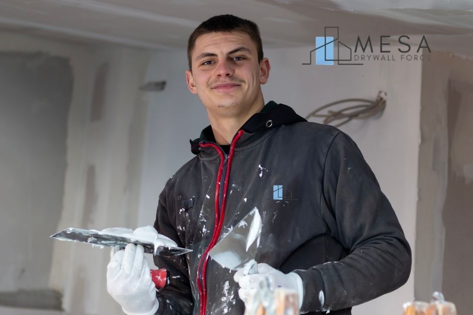 A drywall repair technician in paint-splattered work clothes holds a putty knife and compound container while standing in a residential project with white walls and ceiling visible in the background near E El Sendero Rd, and N Ridgeway Dr in Carefree, AZ.