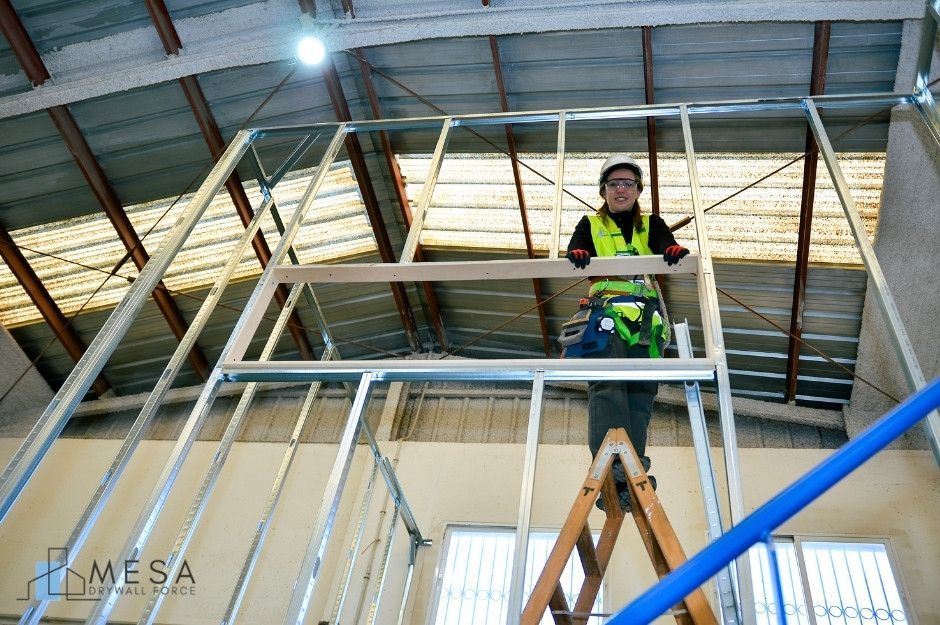 A drywall repair technician, wearing a high-visibility safety vest and hard hat, stands on a wooden stepladder to inspect the metal ceiling framing at a residential construction site. She grips the aluminum framework while examining the drywall structure overhead near W Kaibab Dr, and S Rosemary Pl, Chandler, AZ.