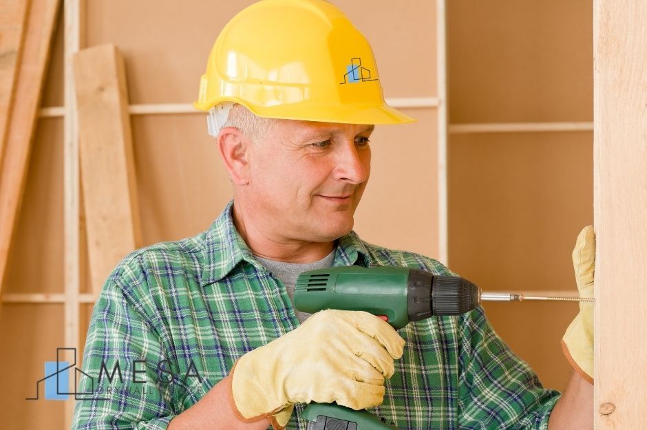 A drywall repair technician in yellow hard hat and green plaid shirt uses a power drill on a commercial construction project with wooden framing and drywall panels visible in the background near W Rancho Ct, and N 185th Ave, Citrus Park, AZ.