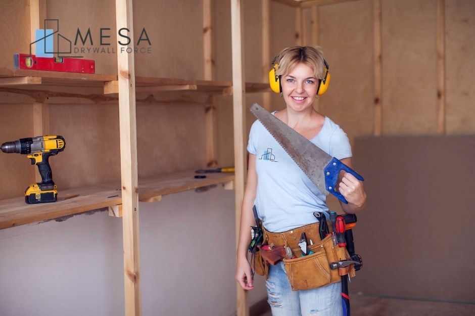 A drywall repair technician wearing a white tank top and tool belt holds a saw while standing in a room with wooden wall framing. She is working on a residential project near W Piute Ave, and N 67th Dr, Glendale, AZ.