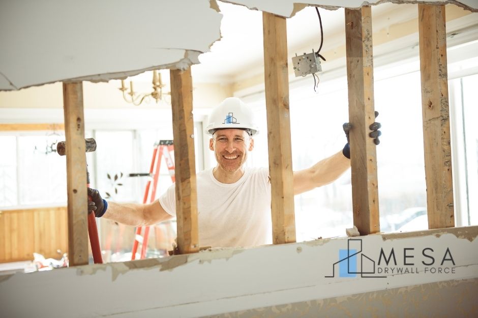 A drywall repair technician in a white hard hat and t-shirt smiles while holding the wooden support beams in a residential construction site with exposed framing and damaged drywall. A step ladder is visible in the background. Located near W Helen Ct, and N Alicia Ct, Maricopa, AZ.