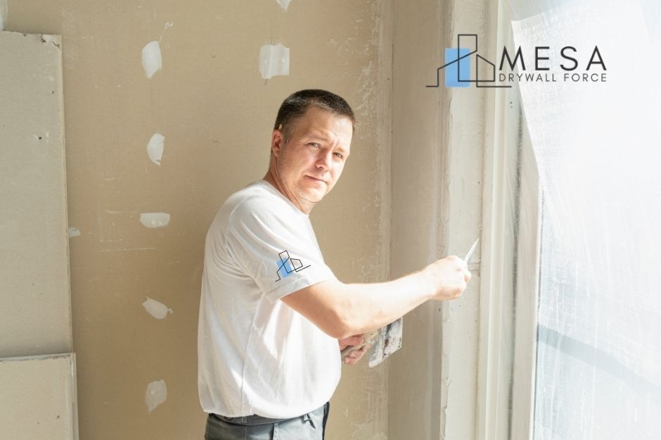 A drywall repair technician applies joint compound using a putty knife during a residential project. He wears a white shirt while working on the unfinished drywall walls, with patch repairs visible throughout the room near E Minnesota Ave, and S Pinewood Dr, Sun Lakes, AZ.