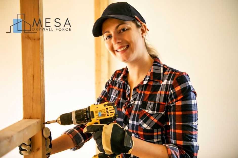 A drywall repair technician in plaid shirt and black cap holds a cordless drill while working on a commercial construction project with wooden framing and white walls visible in the background near W Vogel Ave, and N 185th Ave in Waddell, AZ.