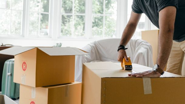 Person sealing a cardboard box with packing tape in a bright room with moving boxes.
