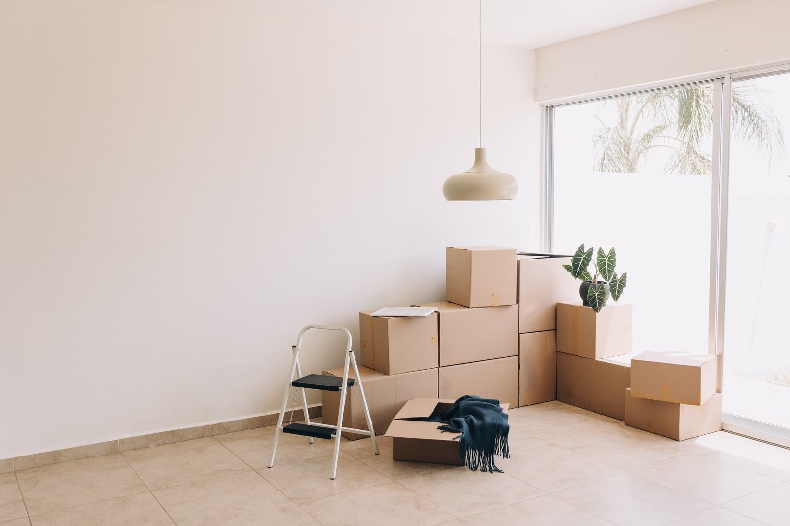 Empty room with moving boxes, a step ladder, and a hanging lamp near a bright window.