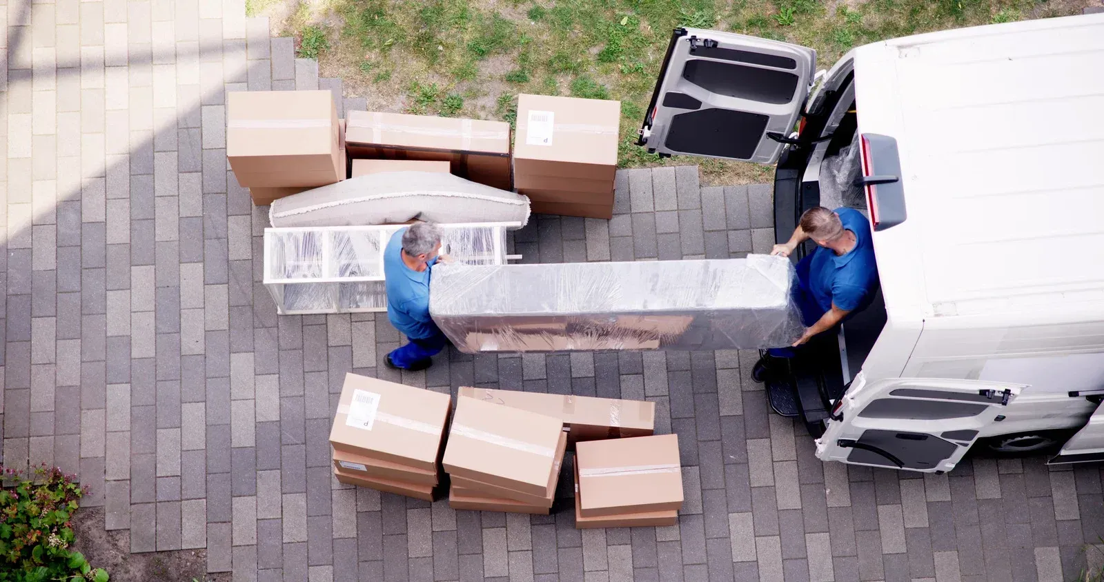 Two movers carry a long white cabinet beside a white van, with cardboard boxes stacked around them.
