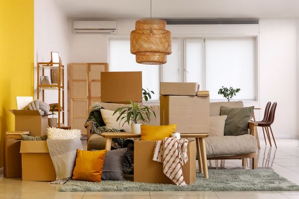 Packed living room with stacked moving boxes, furniture, and a yellow accent chair in a bright apartment