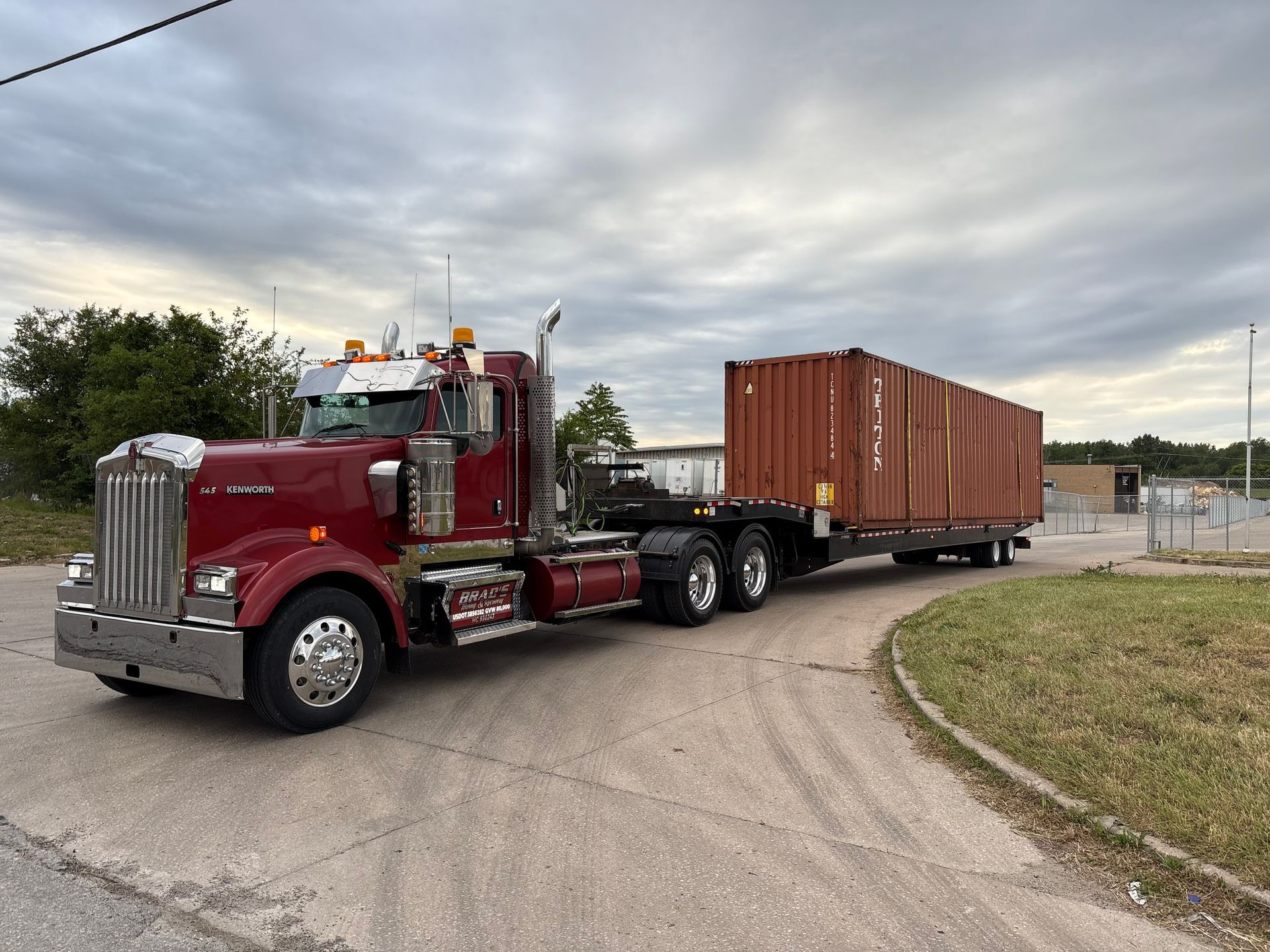 Red semi-truck hauling a brown shipping container on a flatbed trailer, driving on a curved road.