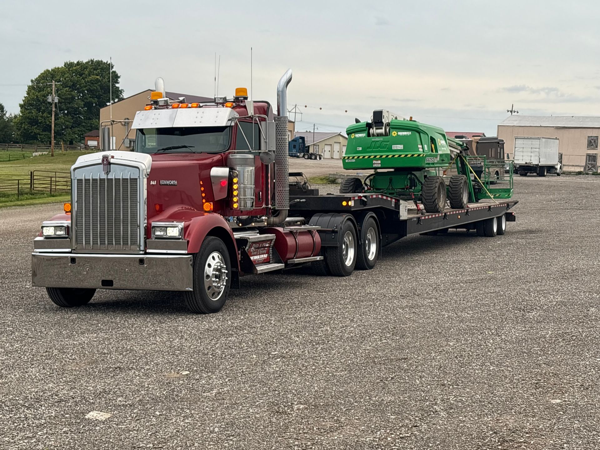 Maroon semi-truck hauling green farm machinery on a flatbed trailer on a gravel lot.