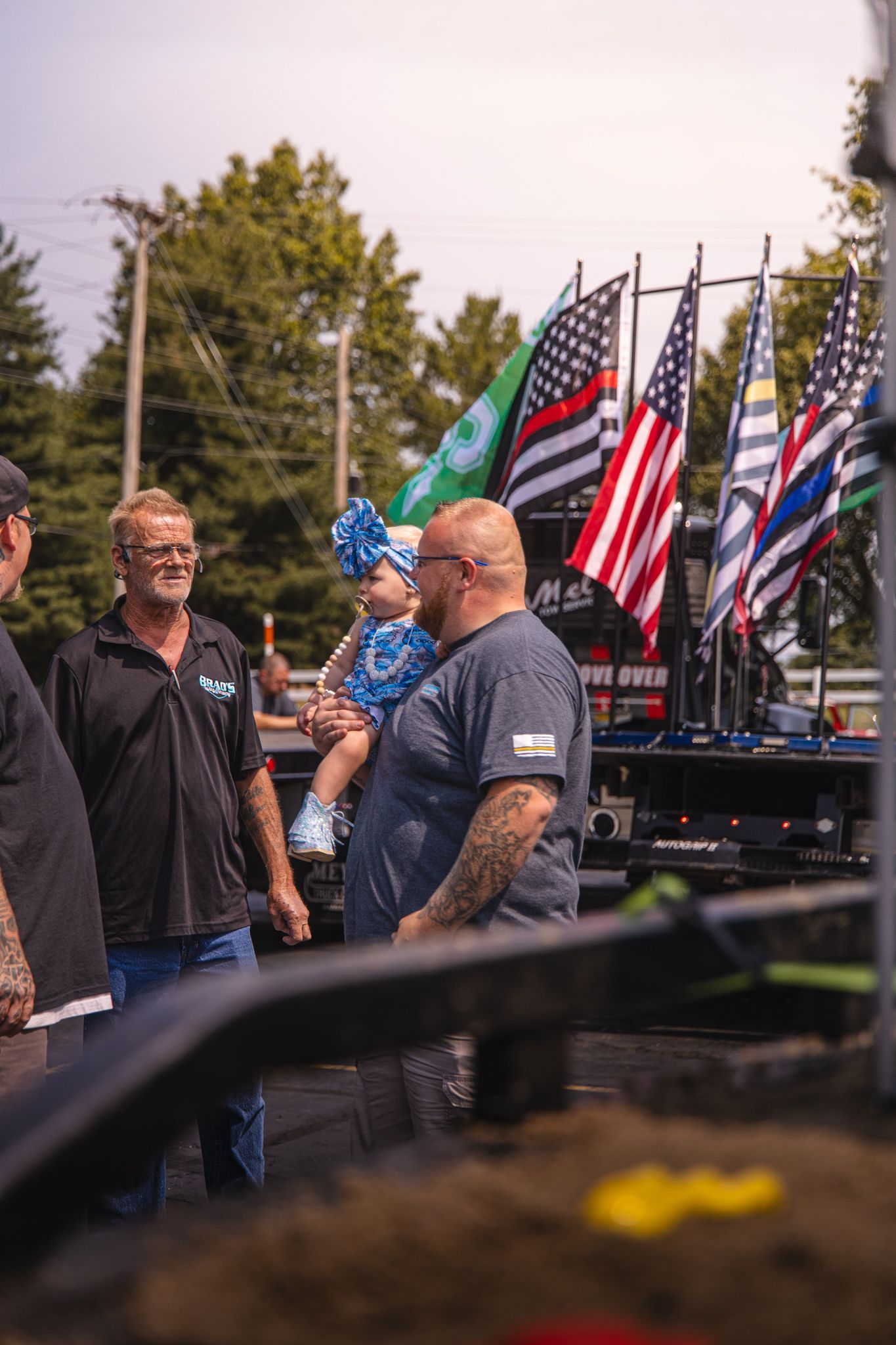 Men and child near flags, one man holds the toddler, the other looks on. Outdoors on a sunny day.