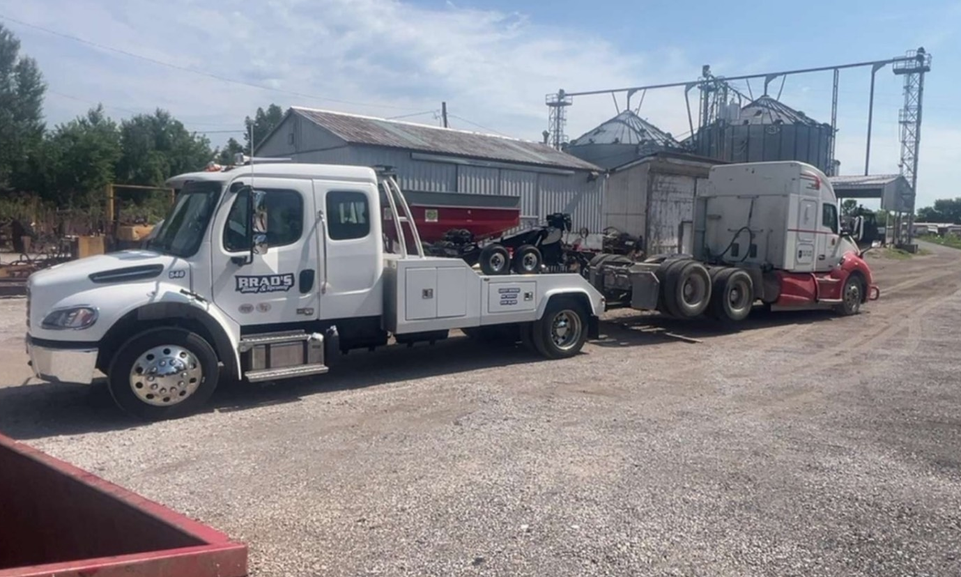 A white tow truck hauls a red and white semi-truck on a gravel lot. In the background are silos and buildings.
