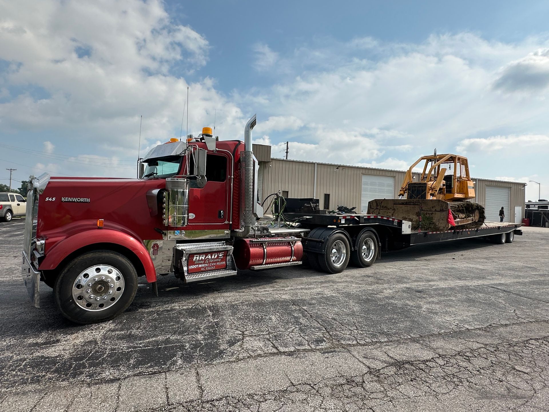 Red semi-truck hauling a yellow bulldozer on a trailer in a parking lot.