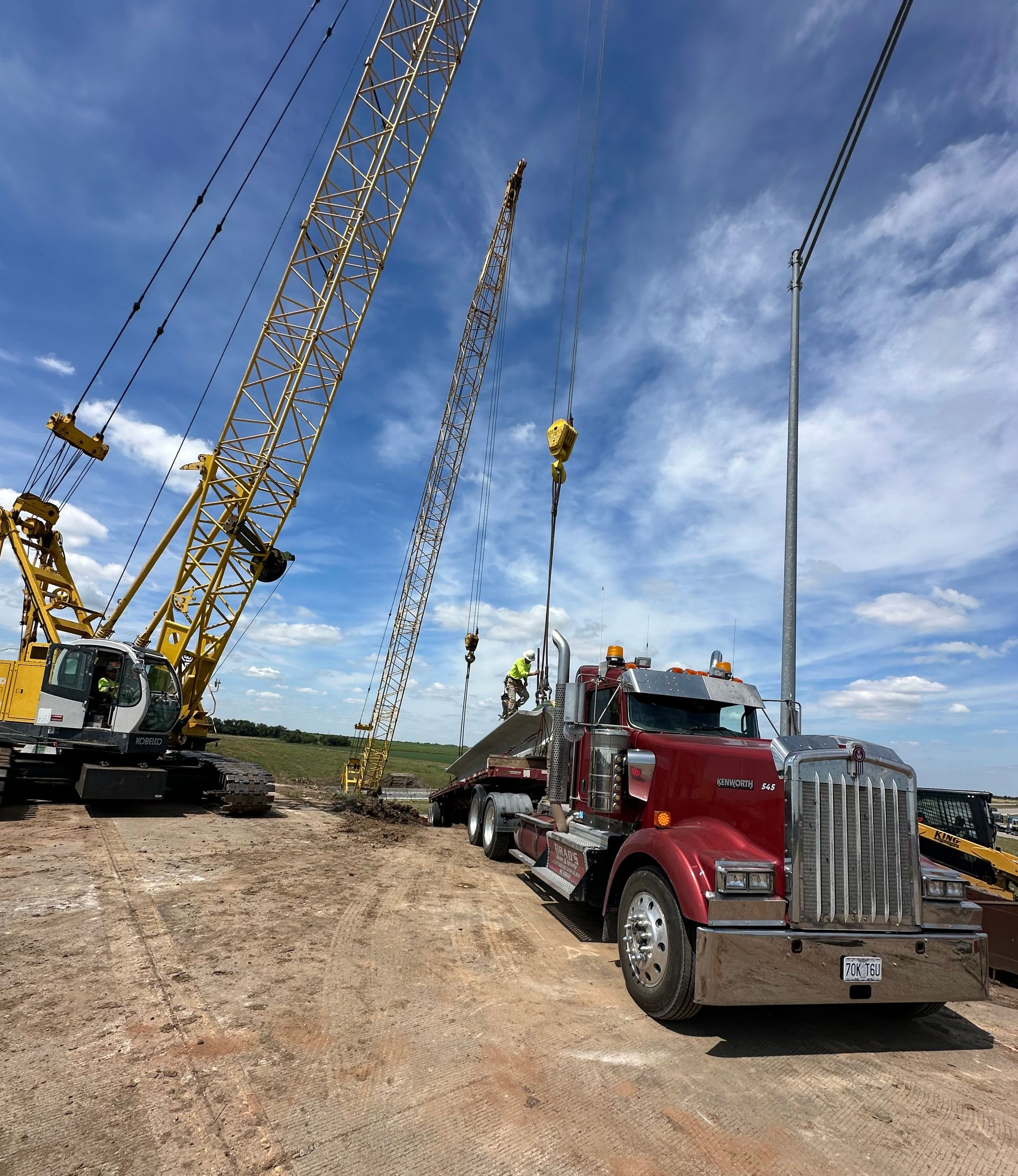 Red semi-truck being loaded by two cranes on a construction site under a partly cloudy sky.