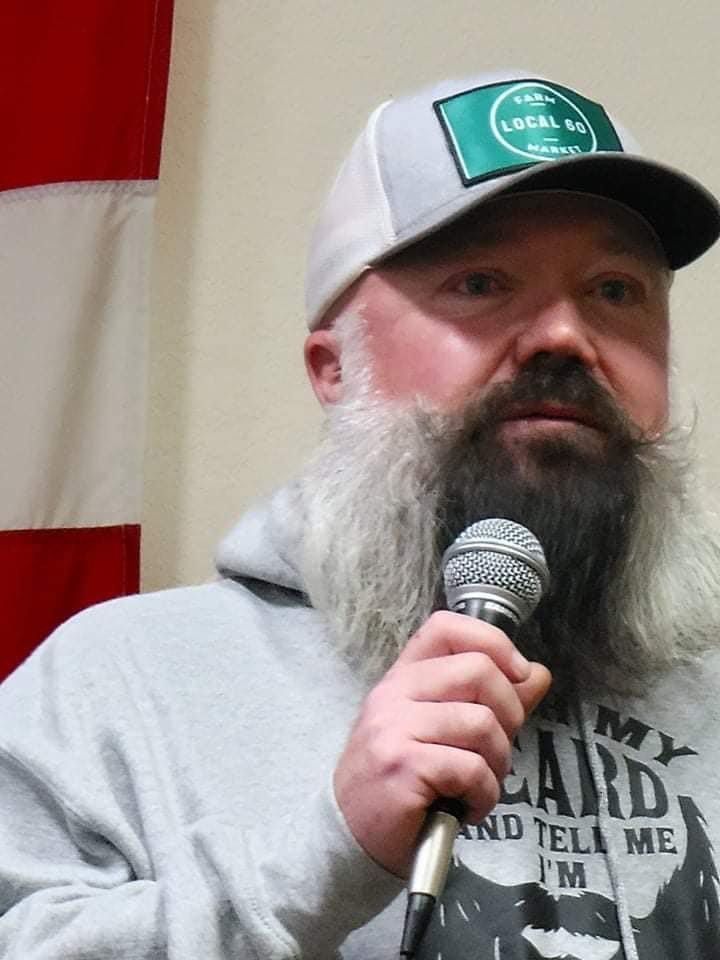 A man with a beard is holding a microphone in front of a flag
