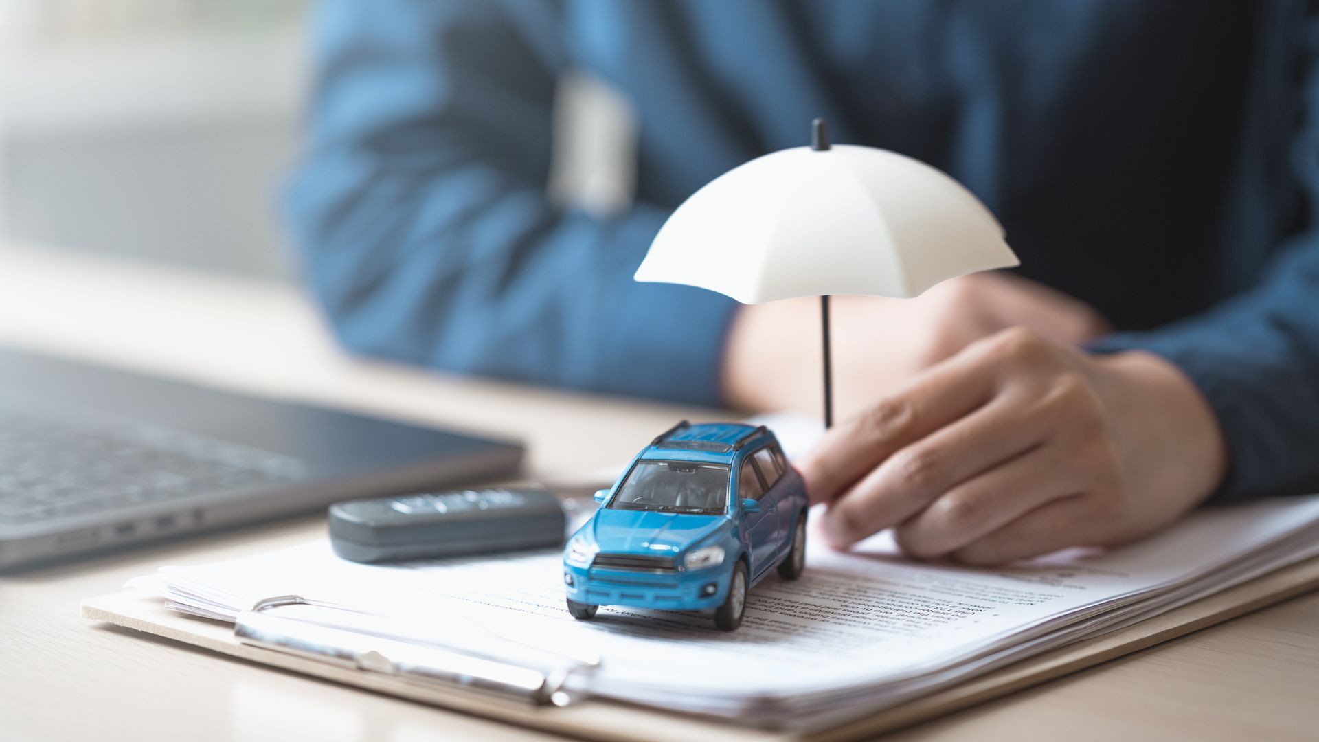 A small car under a miniature umbrella, on top of documents, next to a laptop and remote.