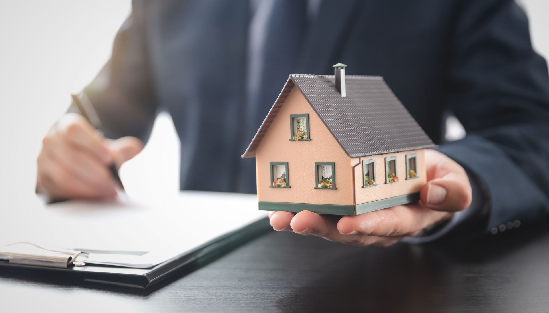 Person in suit holds a miniature house, signing documents on a desk.