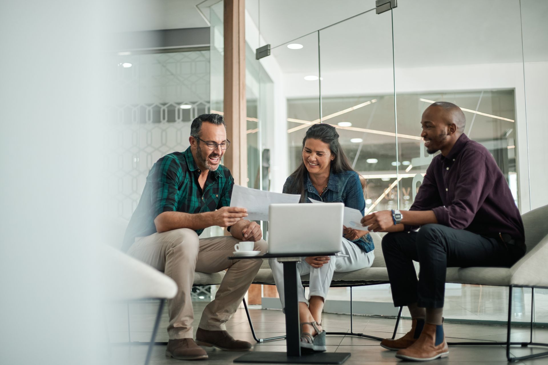 Three people reviewing documents and laptop in a modern office lounge area.