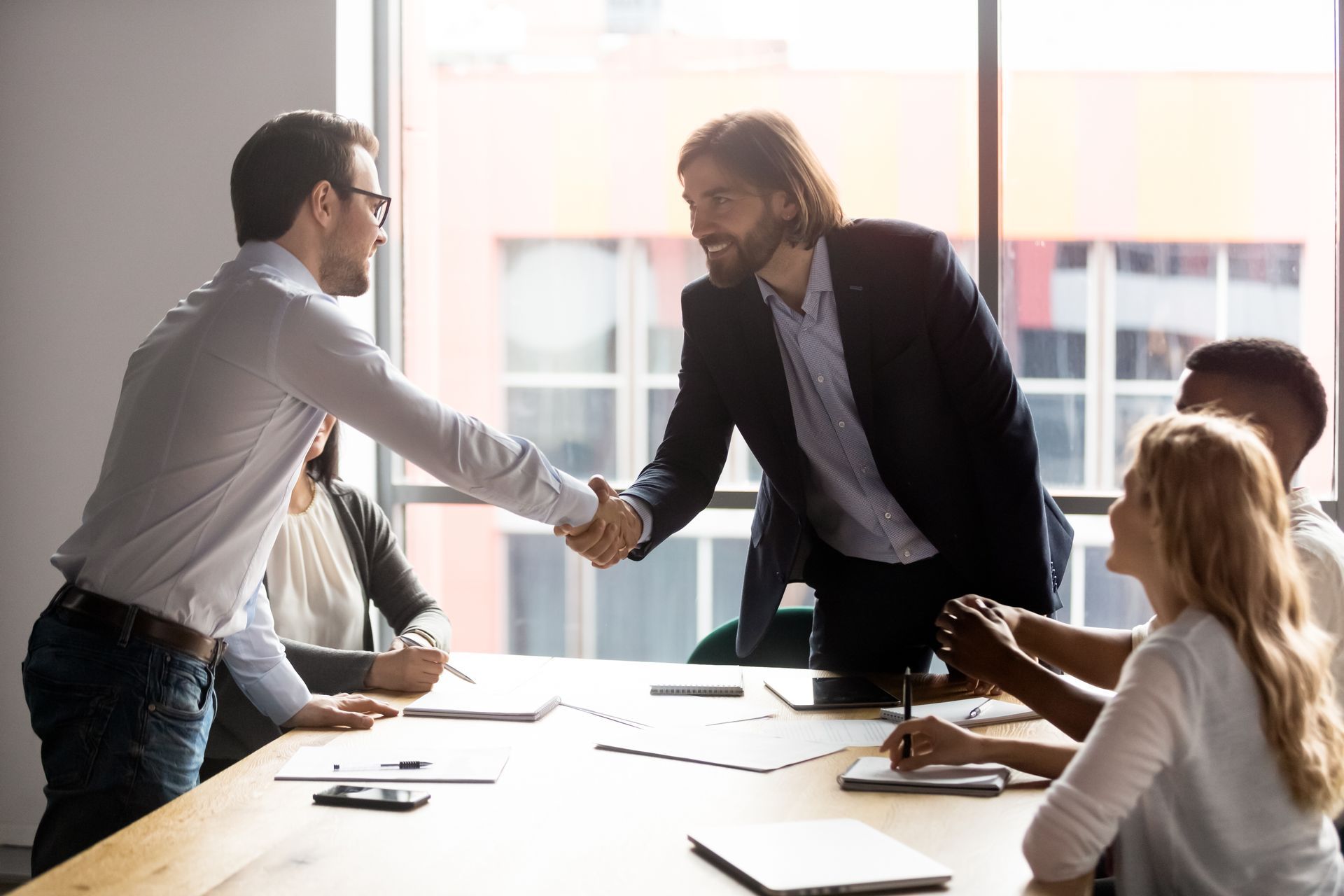 Two men shaking hands across a conference table, other people present in meeting.