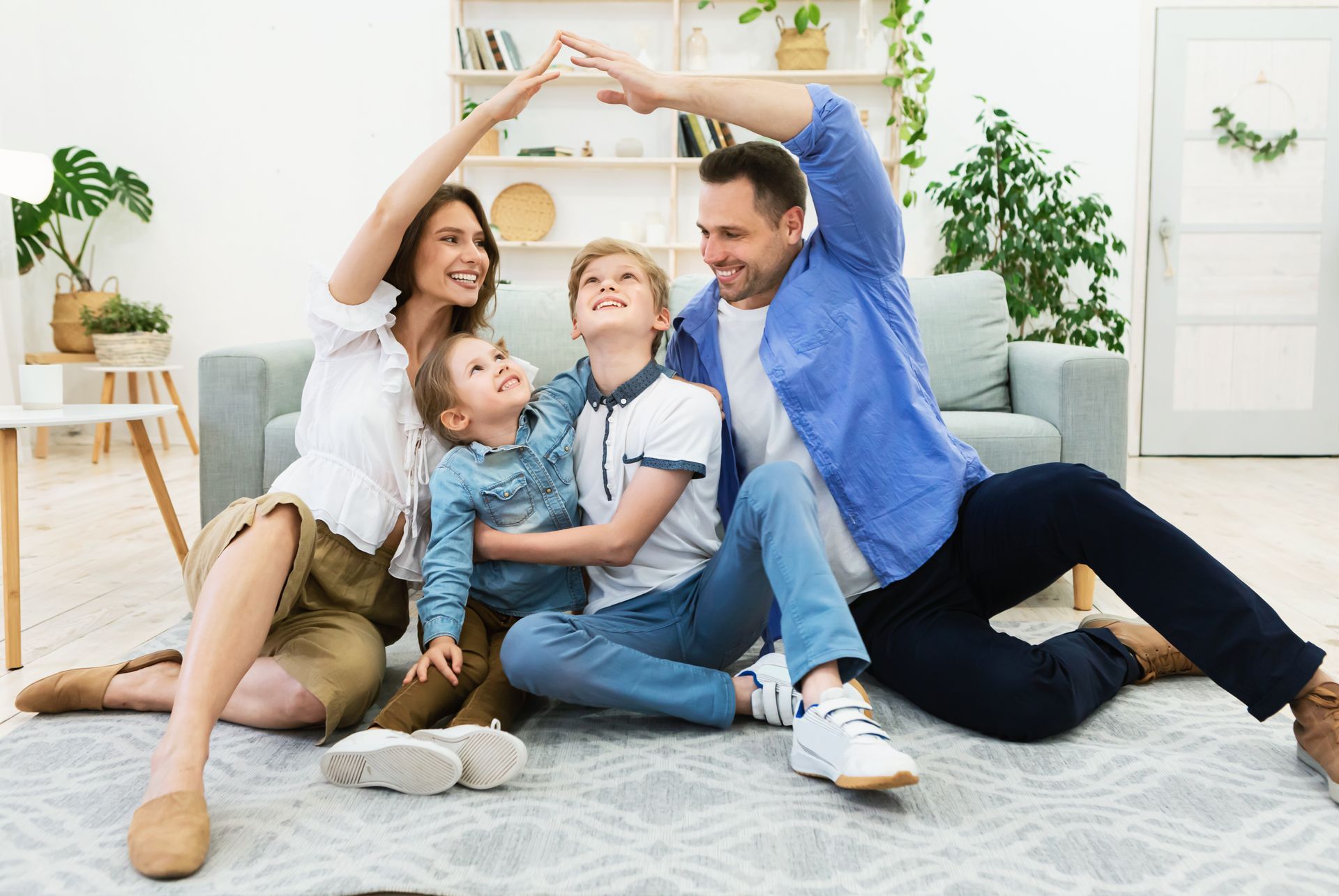 Family sitting on rug, parents making a roof with their hands over two children; light and bright living room.