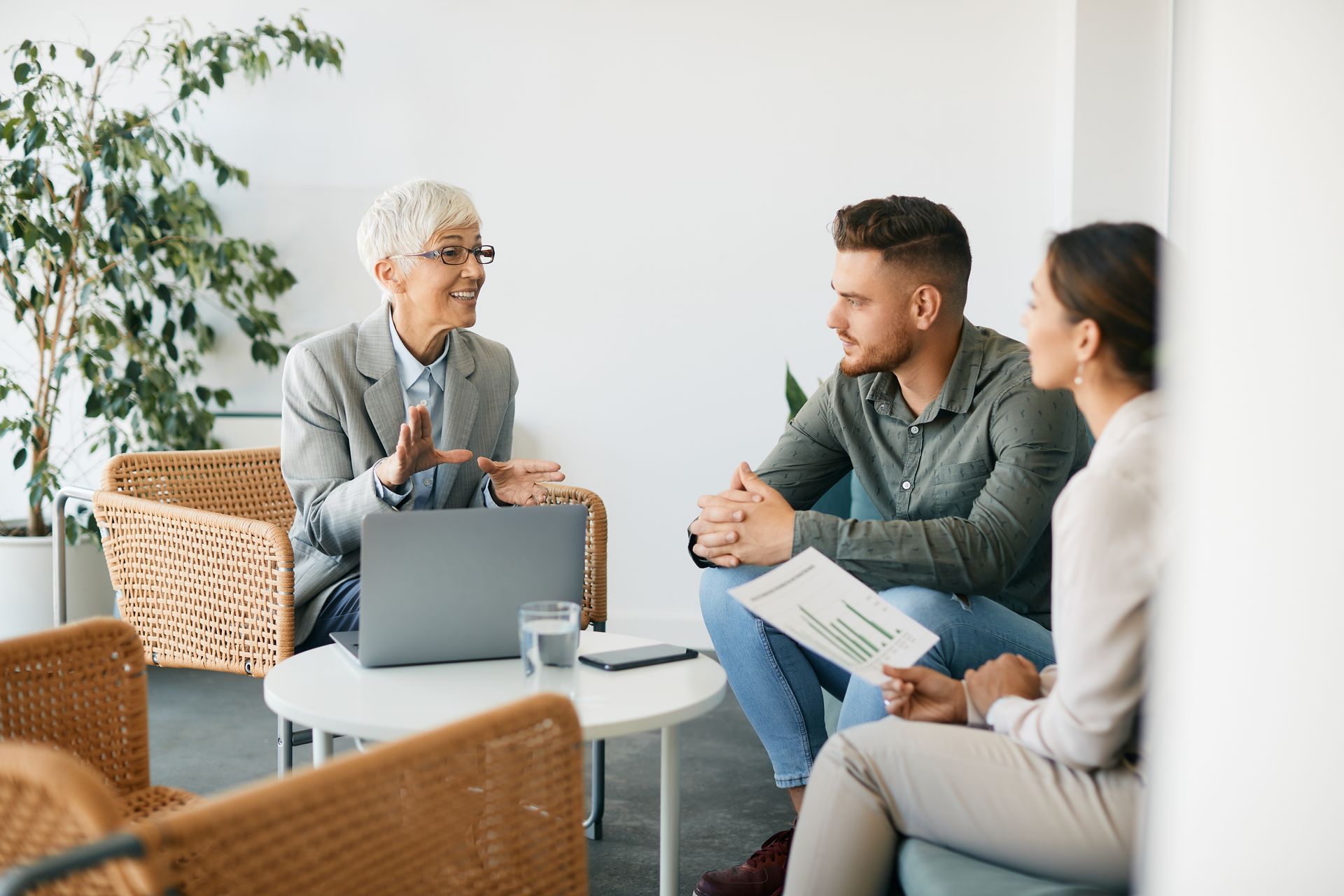 Woman with laptop talking to a couple in a bright office; couple holds a document.