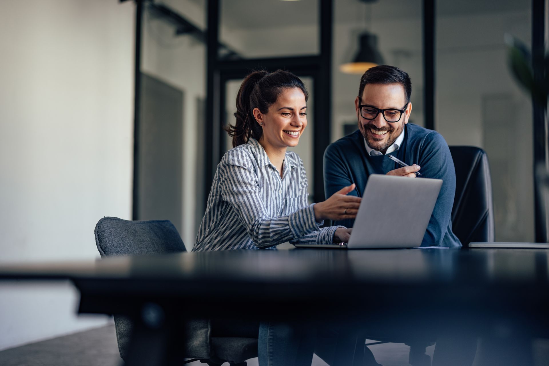 Two people looking at a laptop, smiling.  They are sitting at a dark table in an office.