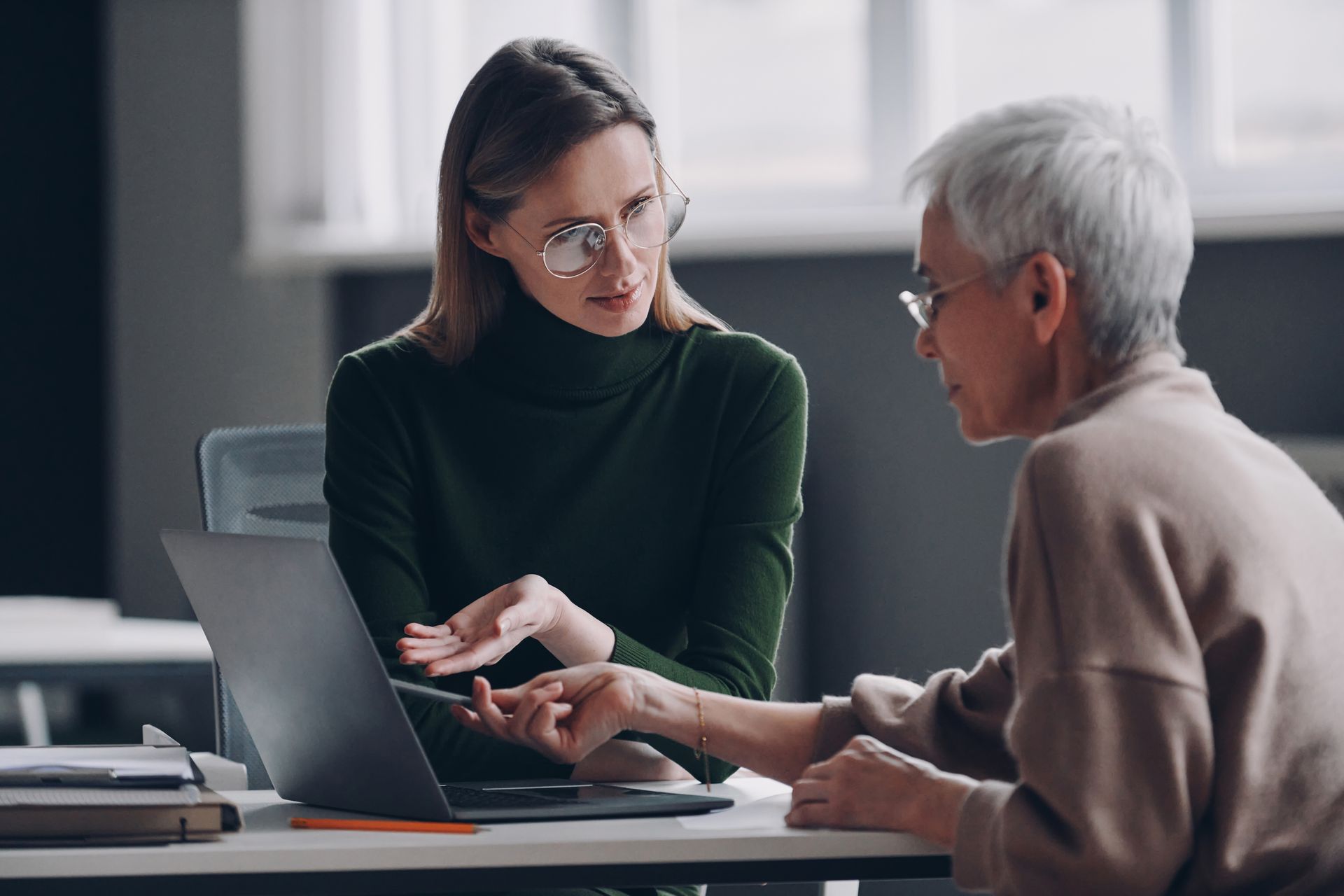 Two people at a desk viewing a laptop. Woman in green turtleneck gestures. Older person points.