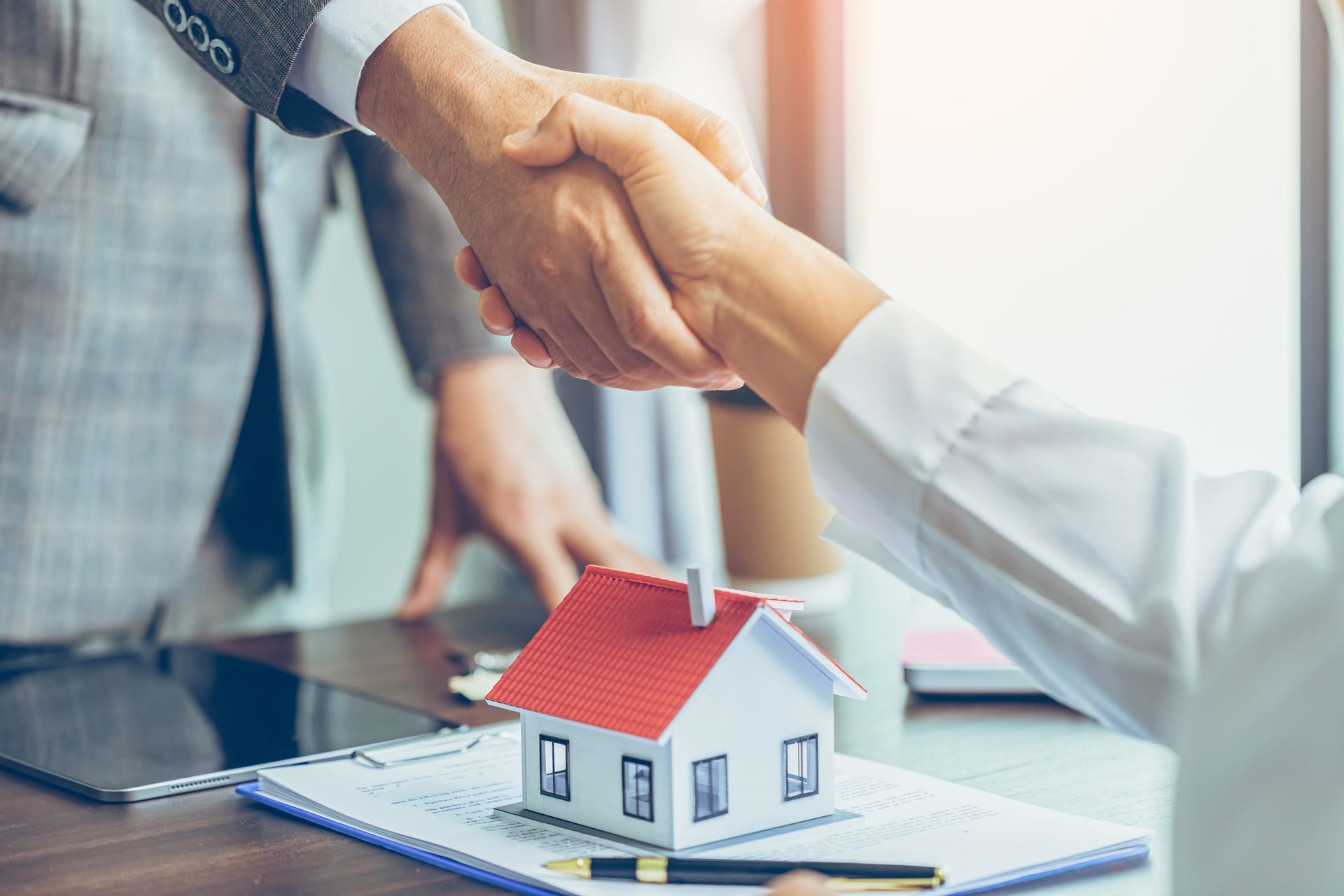 People shaking hands over a miniature house, paperwork, and pen on a desk. Sunlight streams in.
