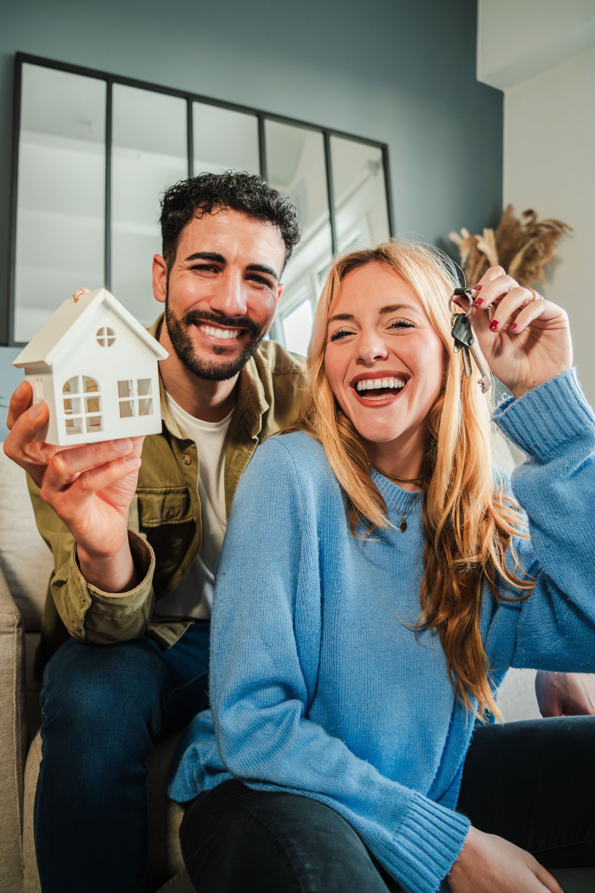Couple smiles, holding house model and keys, celebrating homeownership in a living room.