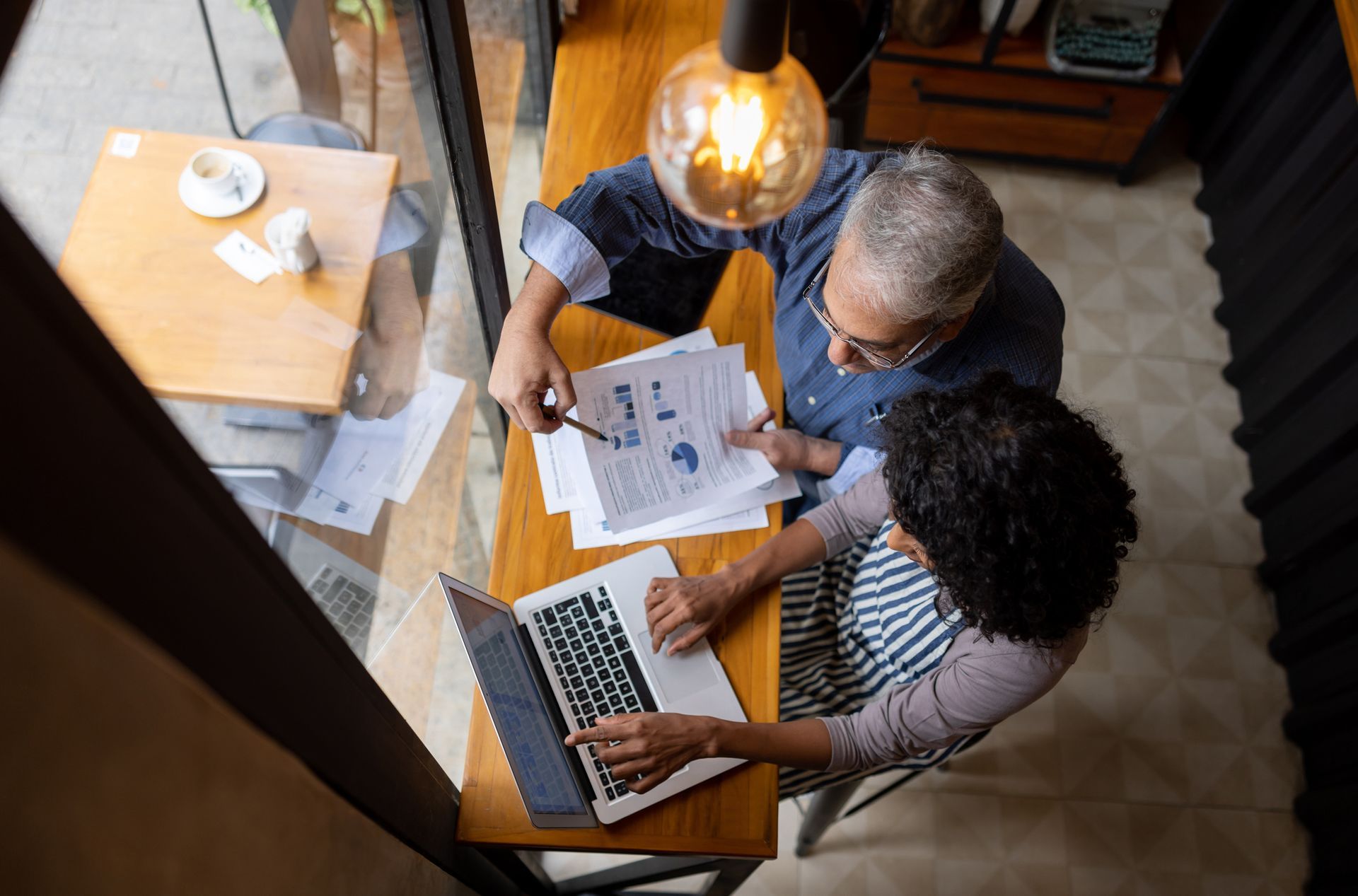 Two people reviewing documents and a laptop at a table, under a hanging light in a cafe.