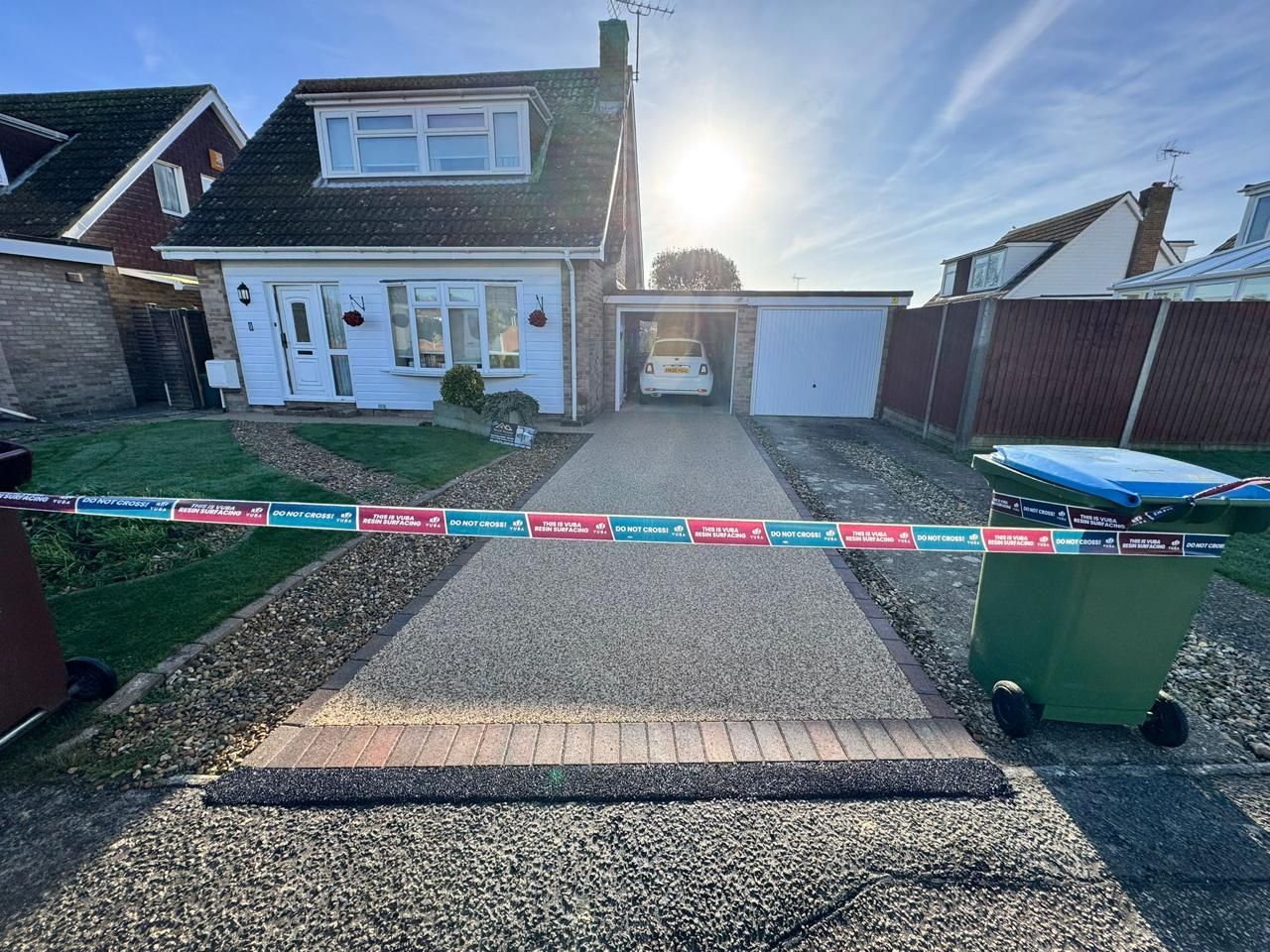 A house with a driveway blocked by police tape. Sunlight shines, and trash cans are present.